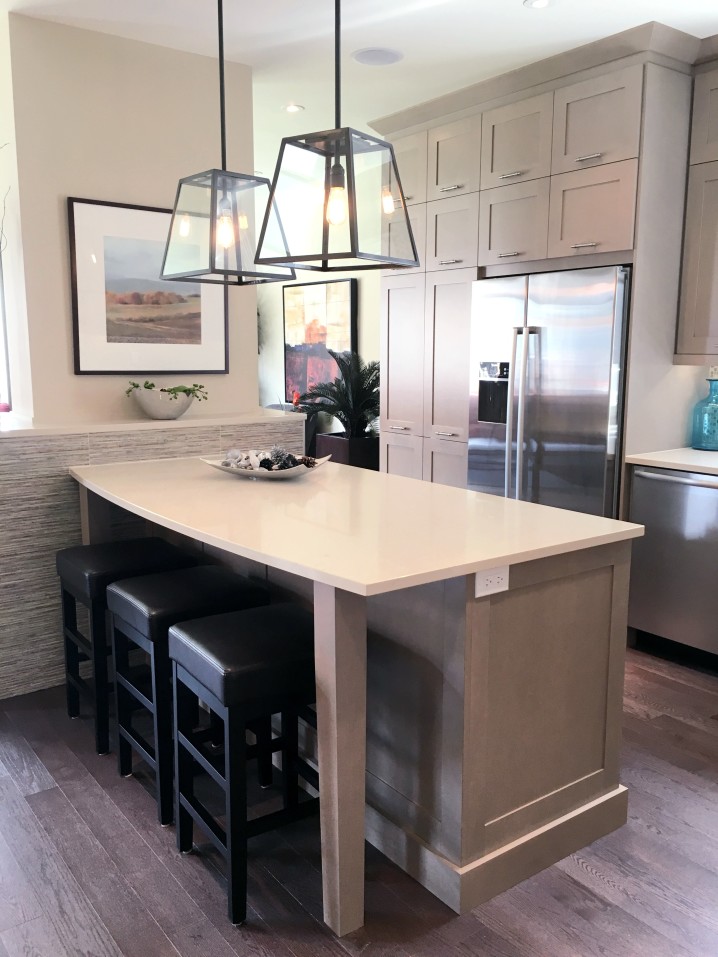 Modern kitchen featuring a quartz island with three black stools, pendant lights, stainless steel appliances, and light wood cabinets. Wall art and decor add a touch of color.