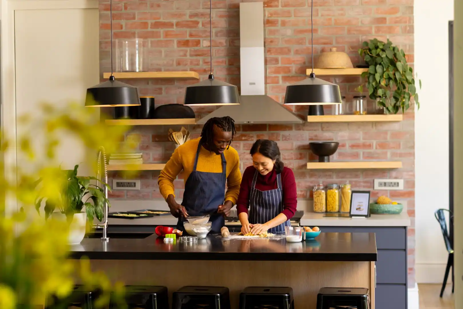 Two people cooking together in a modern kitchen with brick walls and open shelves.