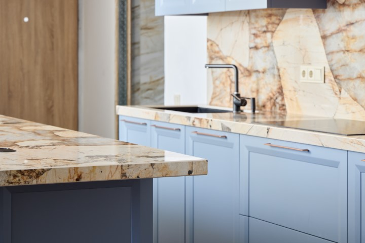 Modern kitchen with light blue cabinets and a marble backsplash, creating a cohesive look. The stone countertops and stainless steel faucet complement the design, while a wood door is visible in the background.