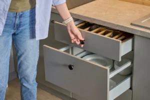 A person is opening a gray kitchen drawer with cutlery, set beneath sleek stone countertops, while another drawer below contains stacked plates.